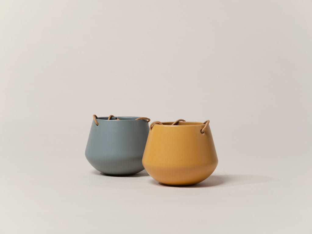 Two spherical planter pots, one in blue-gray and one in tan color, with a matte finish. Each has a small protrusion at the top, likely for hanging, and an adjustable leather cord attached.
